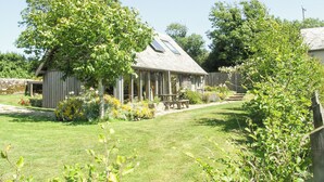 A rustic wooden house with a sloped roof, a covered porch, and a small garden area with a bench. - 2 bedroom accommodation in Holbeton (Holbeton)