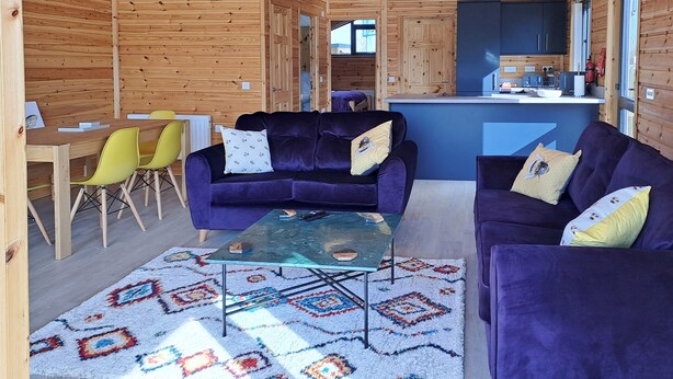 Interior de una cabaña de madera con dos sofás morados, una mesa de centro de cristal, una mesa de comedor con sillas y una zona de cocina.