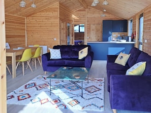 A wooden cabin interior with two purple sofas, a glass coffee table, a dining table with chairs, and a kitchen area. - 2 bedroom accommodation in Brundall (Brundall)