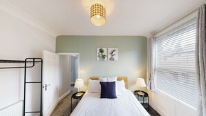 Quarto arrumado com uma cama, luminárias laterais, um radiador, uma janela com persianas e uma porta que dá para outro cômodo.