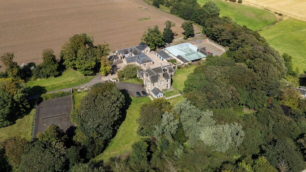 Una vista aerea di un'ampia tenuta con diversi edifici, tra cui una villa principale con un tetto imponente e varie costruzioni più piccole. Si estendono campi verdi e scorre un fiume sullo sfondo.