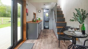 A modern kitchen with a central island, a dining area with a table set for two, and a staircase leading to an upper level. - Little Dunley Cottages - Tiny House (Bovey Tracey, near Newton Abbot)
