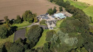 An aerial view of a large estate with multiple buildings, including a main house with a large roof and several smaller structures. There are expansive fields surrounding the property, and a body of water is visible in the background.
