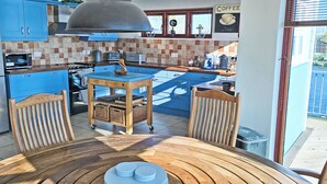 A modern kitchen with blue cabinets, a wooden table, and a view of the outside. - Flag House - UKC7074 (Brundall)