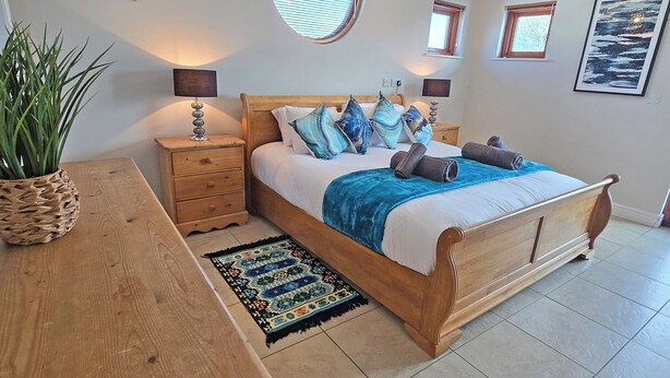 A bedroom with a wooden bed, bedside tables, a nightstand, a potted plant, and a window with blinds. - Flag House - UKC7074 (Brundall)