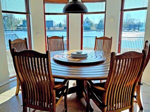 A dining area with a round table and six wooden chairs, large windows with a view of a body of water and buildings, and a black pendant light above the table. - Flag House - UKC7074 (Brundall)