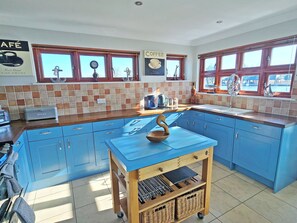 A kitchen with blue cabinets, a wooden table, a coffee station, and a view of the outside. - Flag House - UKC7074 (Brundall)