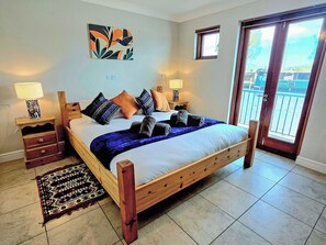 Quarto com cama de madeira, duas mesas de cabeceira, um criado-mudo, tapete estampado e vista para o exterior por uma porta.