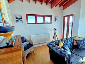 Quarto com piso de madeira, sofá azul, cadeira de vime, modelo de navio e radiador.