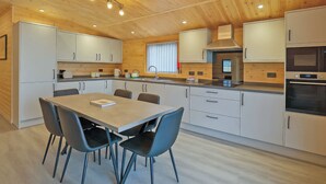A modern kitchen with wooden ceiling, white cabinets, a dining table with chairs, and built-in appliances. - Ferry Lodge - UKC7073 (Brundall)