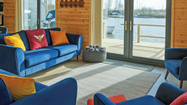 A modern living room with a blue sofa, a grey bean bag, and a view of a dock with boats. - Ferry Lodge - UKC7073 (Brundall)