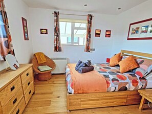 A bedroom with a wooden bed, a chair, a dresser, a window with curtains, and wall art. - Dinky Daze - UKC7071 (Brundall)