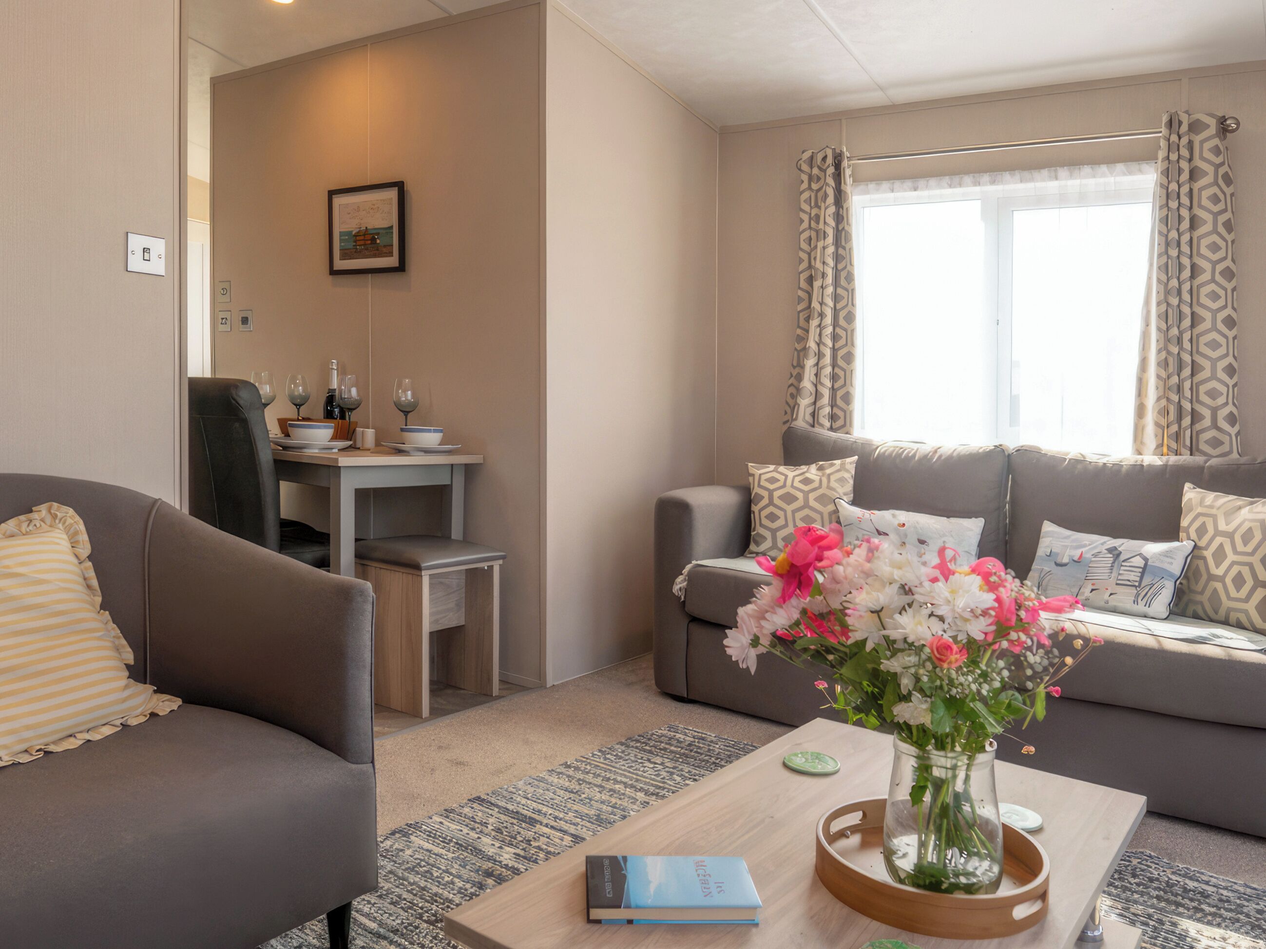 En moderne stue med en sofa, et sofabord med en bog, en vase med blomster, og et spiseområde med et bord dækket til to.