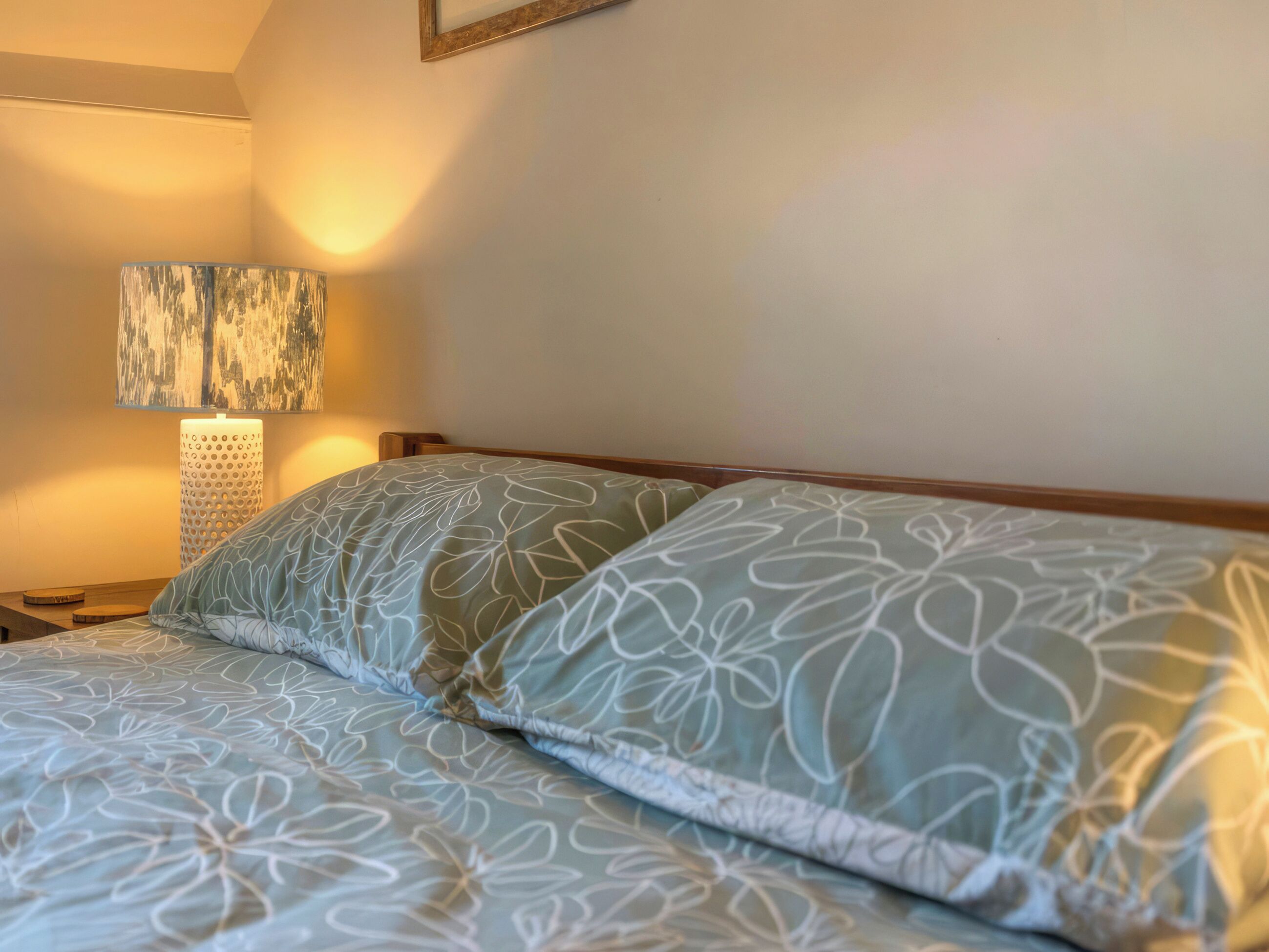 A neatly made bed with floral-patterned bedding, a wall-mounted lamp, and a framed artwork above the bed.