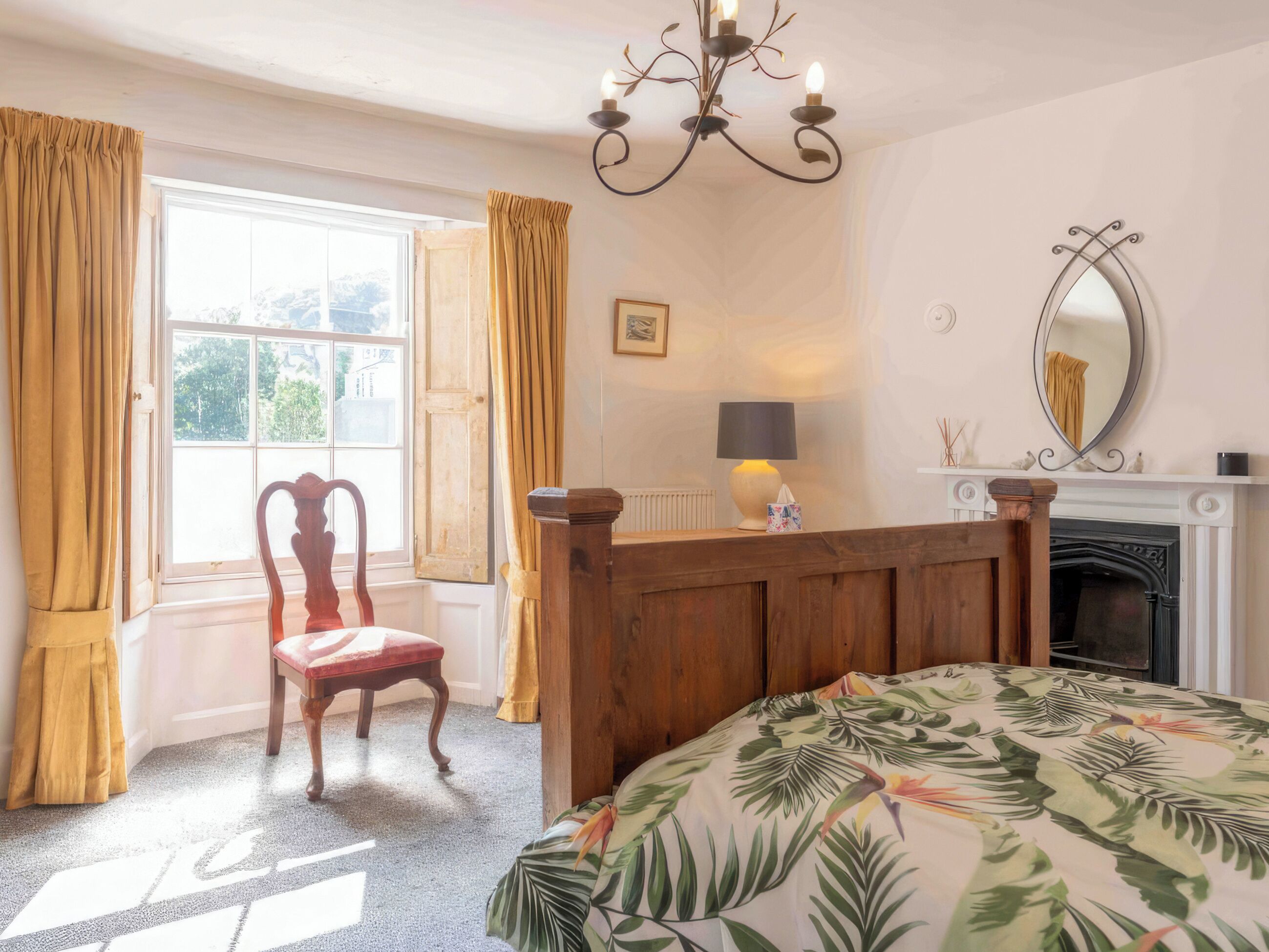 A bedroom with a wooden bed, a chair, a window with curtains, a fireplace, and a chandelier.