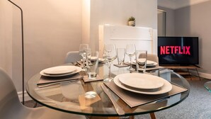 Ein moderner Essbereich mit einem Glastisch, gedeckt für vier Personen, einem Fernseher mit dem Netflix-Logo und einer kleinen Pflanze auf einem weißen Schrank.
