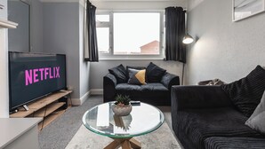 Uma sala de estar moderna com uma grande televisão de tela plana exibindo "NETFLIX", um sofá preto, uma mesa de centro de vidro e uma janela com cortinas.