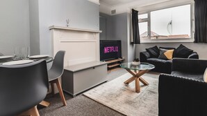 Uma sala de estar moderna com uma TV exibindo 'NETFLIX', uma área de jantar com mesa e cadeiras, e um espaço de estar acolhedor com um sofá e uma mesinha de centro.