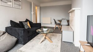 Uma sala de estar moderna com um sofá escuro, uma mesa de centro de vidro e uma televisão de tela plana. Há uma área de jantar com mesa e cadeiras ao fundo.