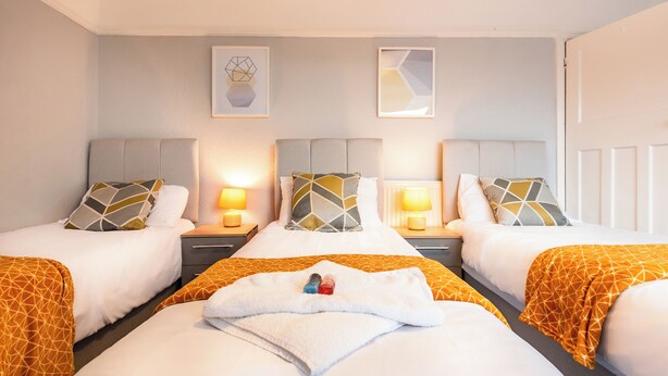 Quarto de hotel com duas camas, cada uma com cabeceira, mesa de cabeceira, abajur e almofada estampada com padrão geométrico.