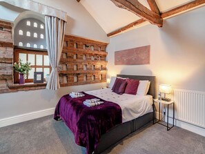 A bedroom with a brick wall, a window with curtains, a bed with a purple blanket, and a bedside table with a lamp. - 1 bedroom accommodation in Bilbrough, near York (Bilbrough, near York)