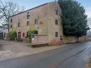 A brick building with a large tree in front, a gravel driveway, and a road to the right.