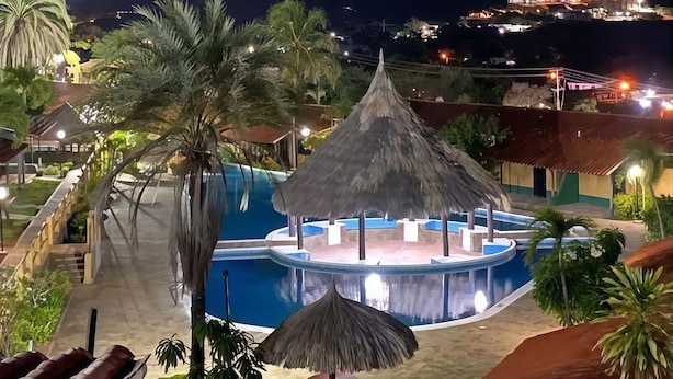 Zona de piscina en un resort con una estructura de techo de paja, palmeras y vistas a montañas distantes.