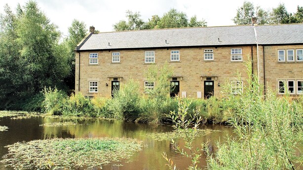 A two-story stone building with multiple windows, a pond in front, and surrounding greenery. - Peveril Cottage - E4642 (Darwin Lake, near Matlock)