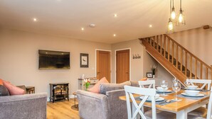 A modern living room with a wooden staircase, a dining area with white chairs, and a fireplace.