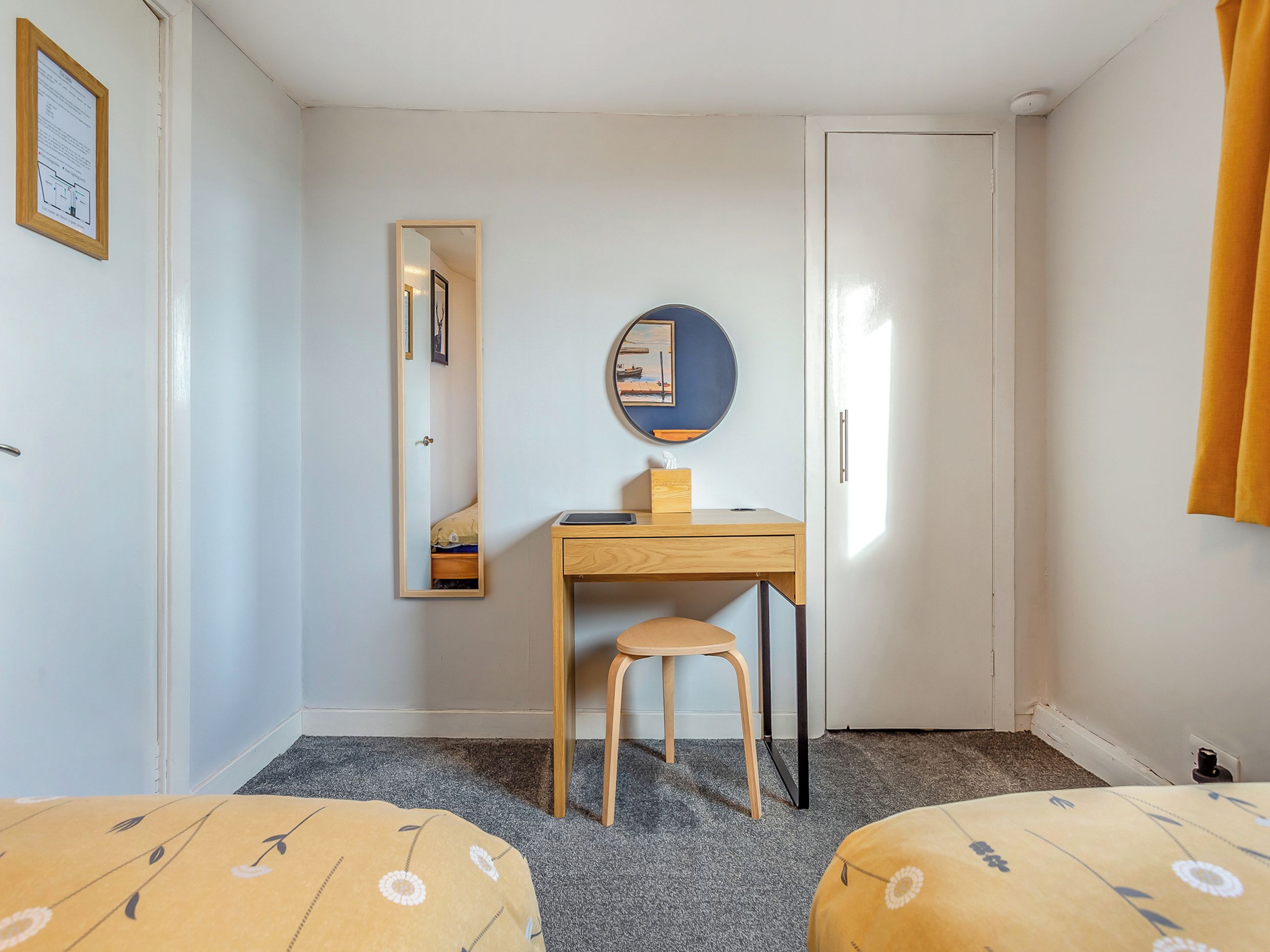 A room with a desk, chair, two beds, a mirror, a door, and a framed notice on the wall.