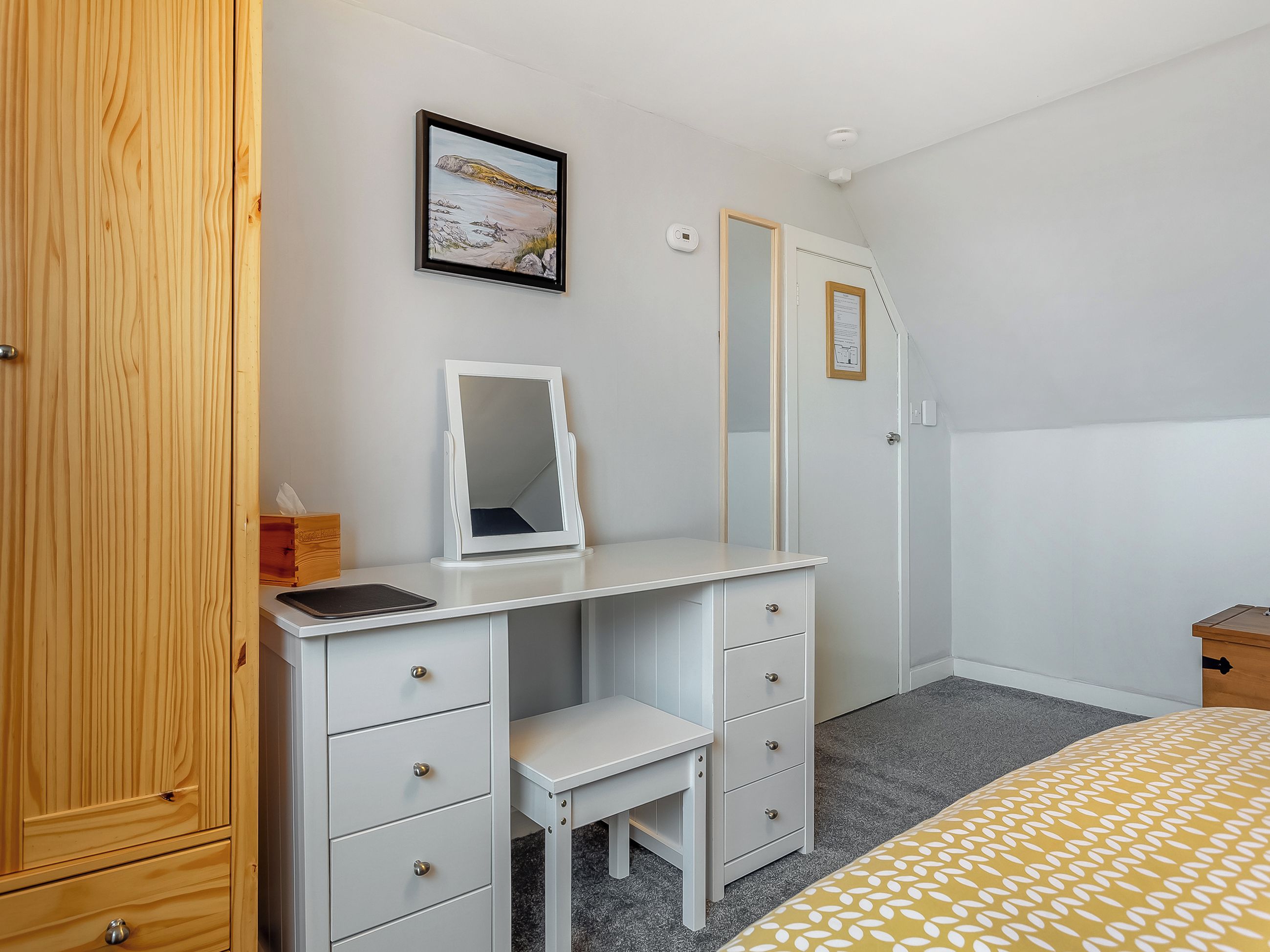 Ein Schlafzimmer mit weißem Schminktisch, einem Spiegel, einem Bett mit gemustertem Bettbezug, einem Holzschrank und einem gerahmten Bild an der Wand.
