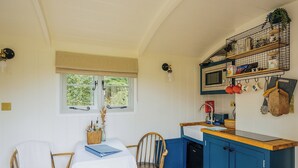 A compact kitchen with a wooden table, two chairs, a window with curtains, and a blue cabinet with a microwave and utensils. - UK41468 - Lapwing Hut (Cholderton, near Salisbury)