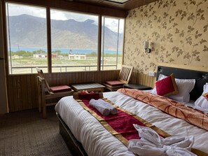 A hotel room with a bed, a wooden chair, a small table, and a view of mountains and a lake. - The Hidden Heaven Cottage Pangong (Leh)