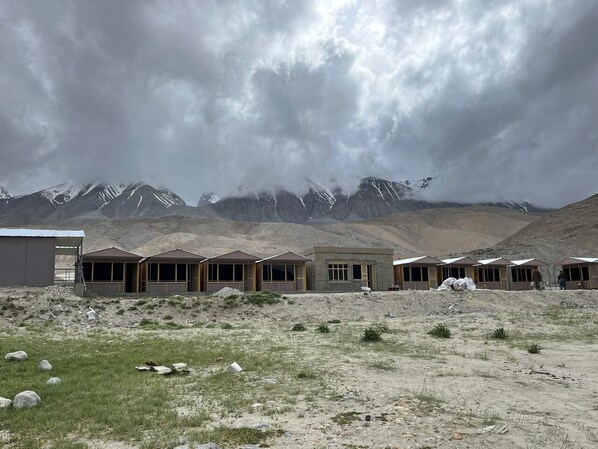 A row of simple, single-story buildings with large windows, set against a backdrop of mountains and a cloudy sky.