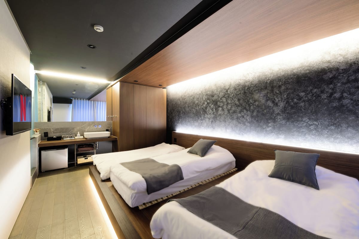 A modern hotel room with two beds, a small kitchenette, and a large mirror.