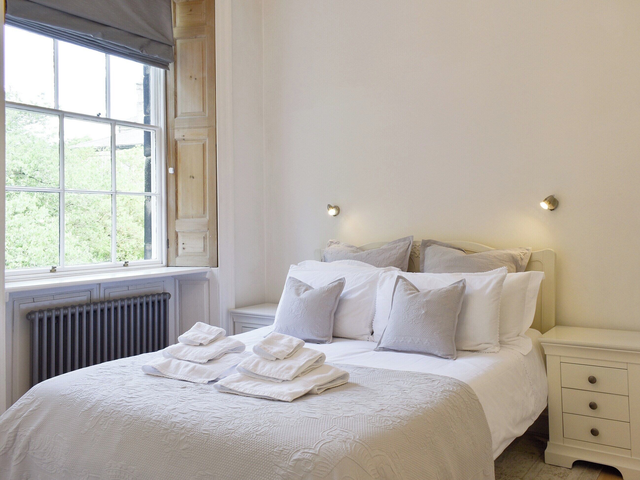 A neatly made bed with folded towels, a nightstand with a lamp, and a window with a view of greenery.