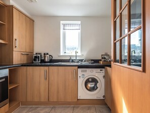 A compact kitchen with wooden cabinets, a built-in washing machine, a coffee maker, and a window providing natural light. - 2 bedroom accommodation in Ashford (Ashford)