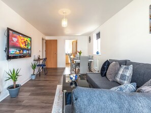 A living room with a flat-screen TV, grey sofa, and a dining area in the background. - 2 bedroom accommodation in Ashford (Ashford)