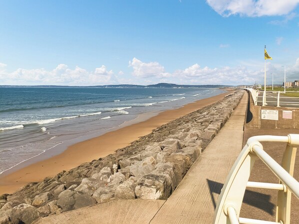 A coastal walkway with a rocky barrier, a sandy beach, and a clear sky.