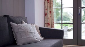 A grey sofa with two white pillows, a window with floral curtains, and a view of greenery outside.