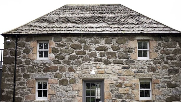 A stone building with a grey door, two windows on the upper floor, and a sloped roof.