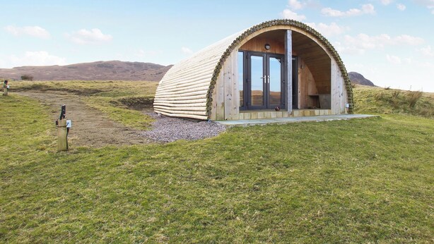 Une structure en bois en forme de dôme, dotée d’une porte et de fenêtres, située dans un champ verdoyant sous un ciel dégagé.