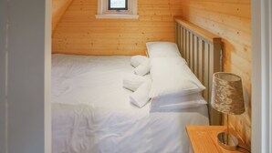 Une chambre avec un plafond en pente en bois, un lit simple avec du linge de lit blanc, une lampe de chevet et une petite fenêtre.