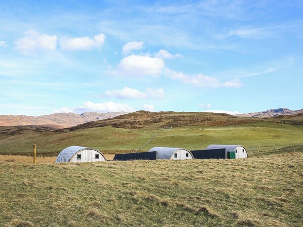 A rural landscape with three small, white, single-story buildings with green roofs, surrounded by open fields and distant mountains under a clear sky.