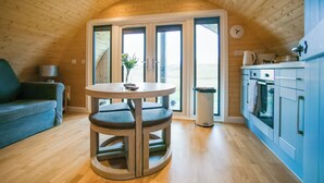 L’intérieur chaleureux d’un chalet comprend une table ronde en bois, un canapé, un coin cuisine et une grande porte-fenêtre coulissante qui donne sur un paysage extérieur magnifique.