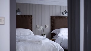 Deux lits avec du linge de lit blanc, des têtes de lit en bois, une petite table sur laquelle est posé un vase de fleurs et des lampes fixées au mur.