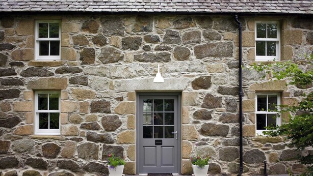 Une maison en pierre avec une porte grise, deux fenêtres de chaque côté et un petit porche agrémenté de plantes en pot.