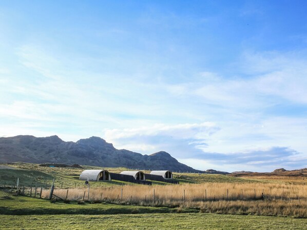 A rural landscape with a series of small, open-roofed buildings, possibly farmhouses, set against a backdrop of rolling hills and mountains. - A good sized, dog friendly pod with silent surroundings. (Archaracle)