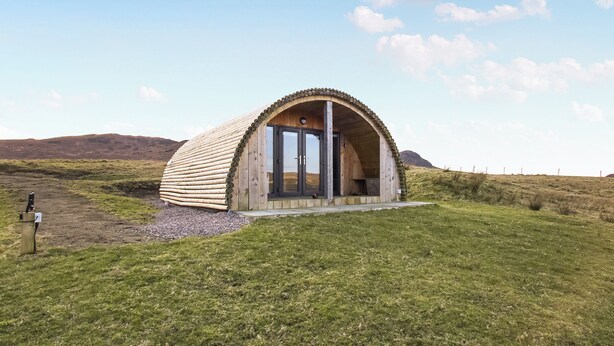 A dome-shaped wooden structure with a door, set in a grassy field with hills in the background. - A good sized, dog friendly pod with silent surroundings. (Archaracle)