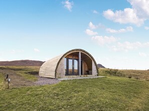 A dome-shaped wooden structure with a door, set in a grassy field with hills in the background. - A good sized, dog friendly pod with silent surroundings. (Archaracle)